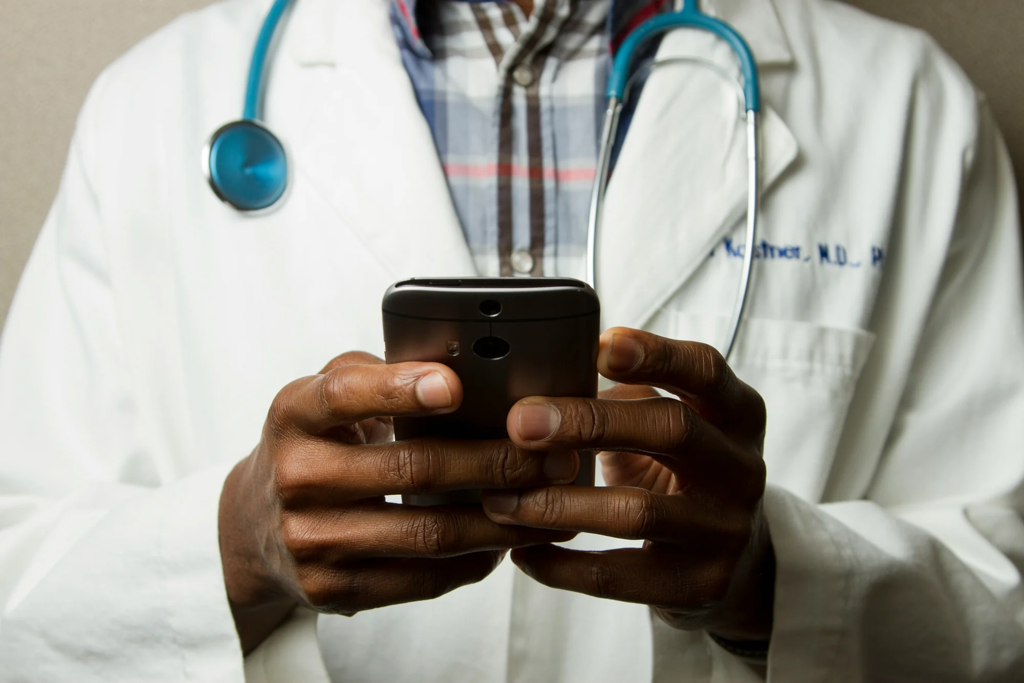 A clinician in a white coat with a stethoscope holds a phone in both hands.